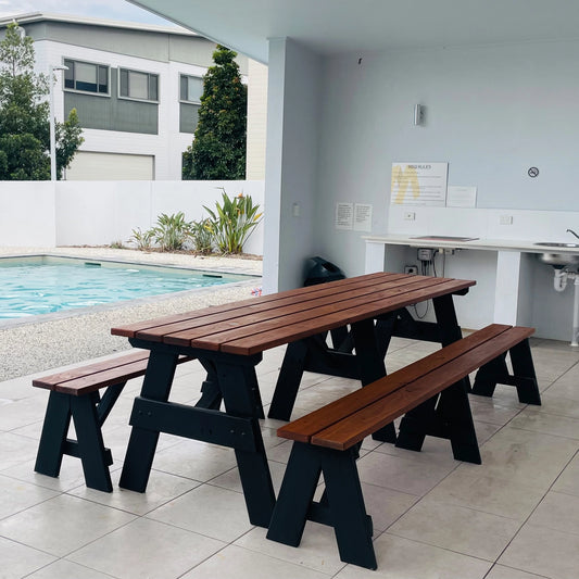 A wooden picnic table with two matching benches, featuring medium brown slatted tops and black wooden A-frame legs, set in an outdoor patio area next to a swimming pool.