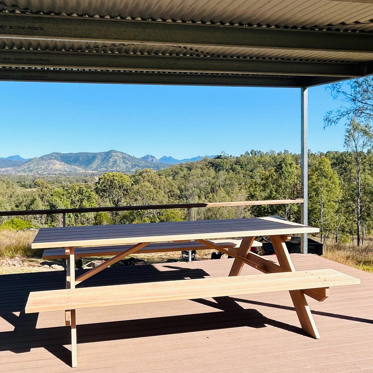 A wooden picnic table with attached benches sits on a wooden deck under a covered patio, with a scenic mountain and forest landscape in the background. Handmade by JJ Timber Design