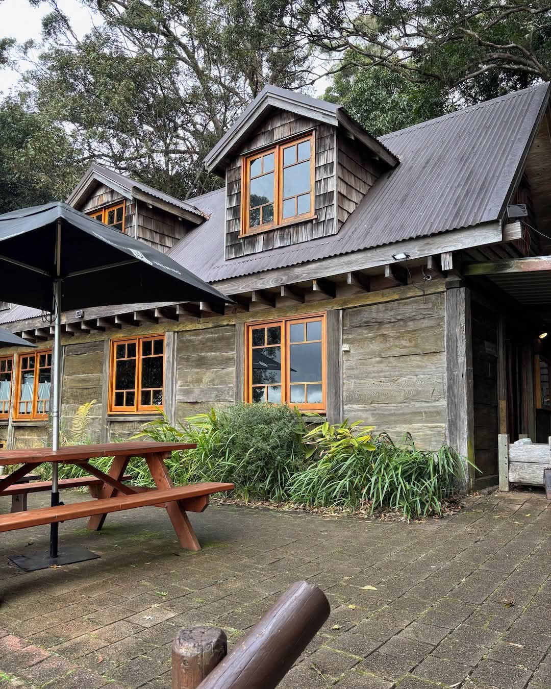A rustic wooden cabin with gray metal roofing, orange-framed windows, and a paved patio area with a picnic table and umbrella, surrounded by greenery and trees.