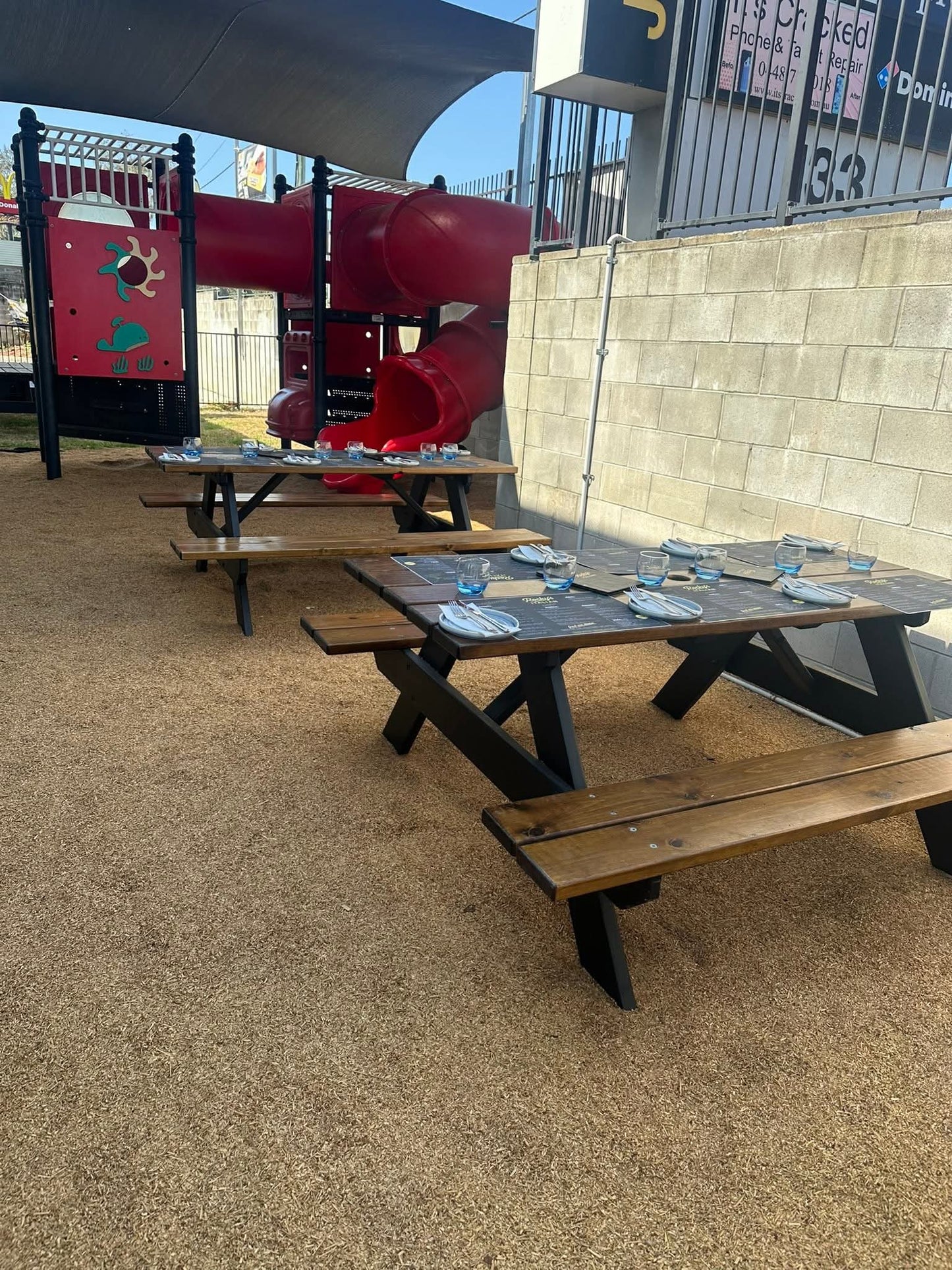 Outdoor wooden picnic tables with attached benches set on a sandy play area beside a red playground structure and a beige block wall.  Wheelchair friendly