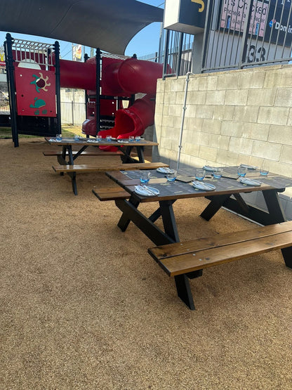 Outdoor wooden picnic tables with attached benches set on a sandy play area beside a red playground structure and a beige block wall.  Wheelchair friendly