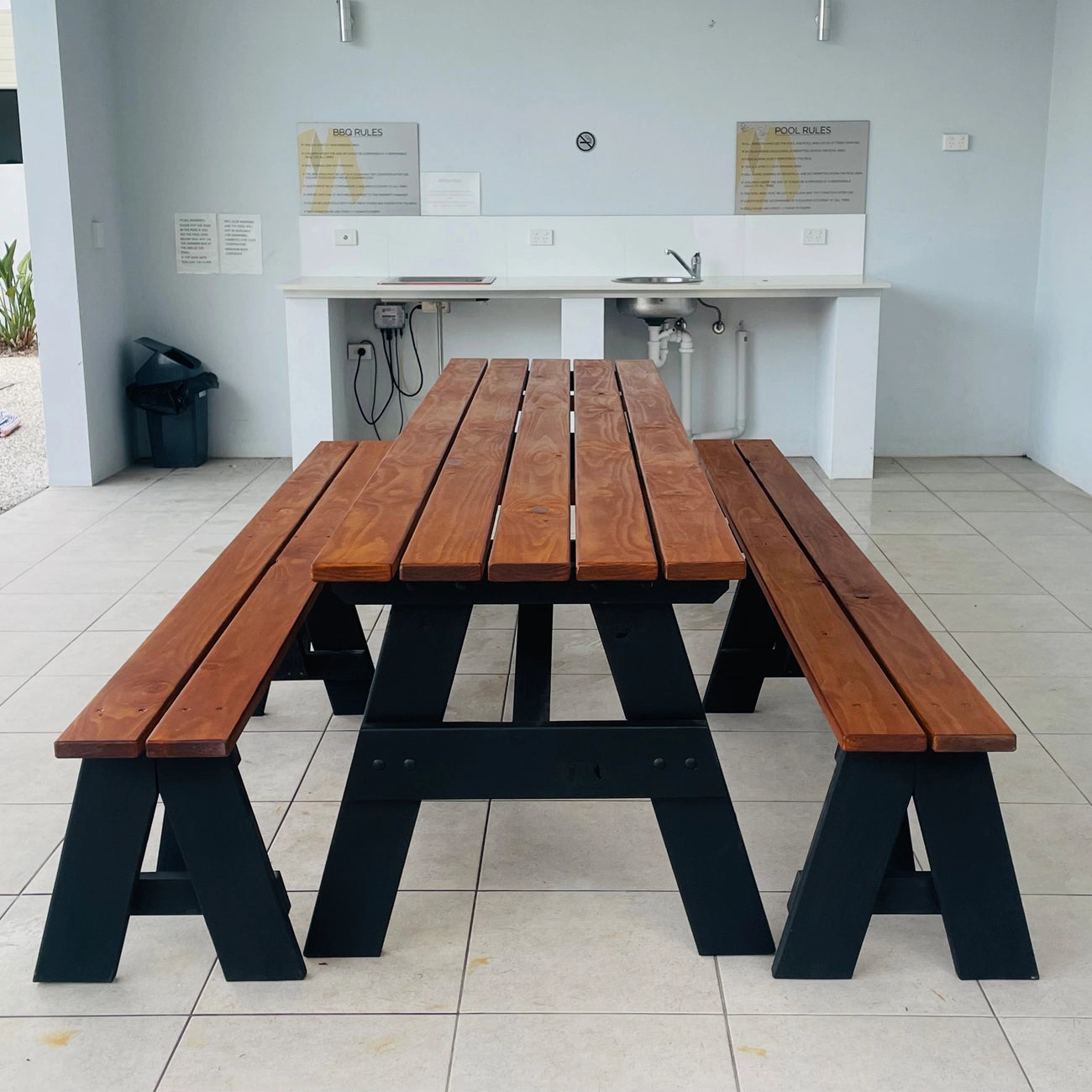 A wooden picnic table with attached benches, featuring medium brown wood slats and black wooden A-frame legs, set in a tiled outdoor patio area.