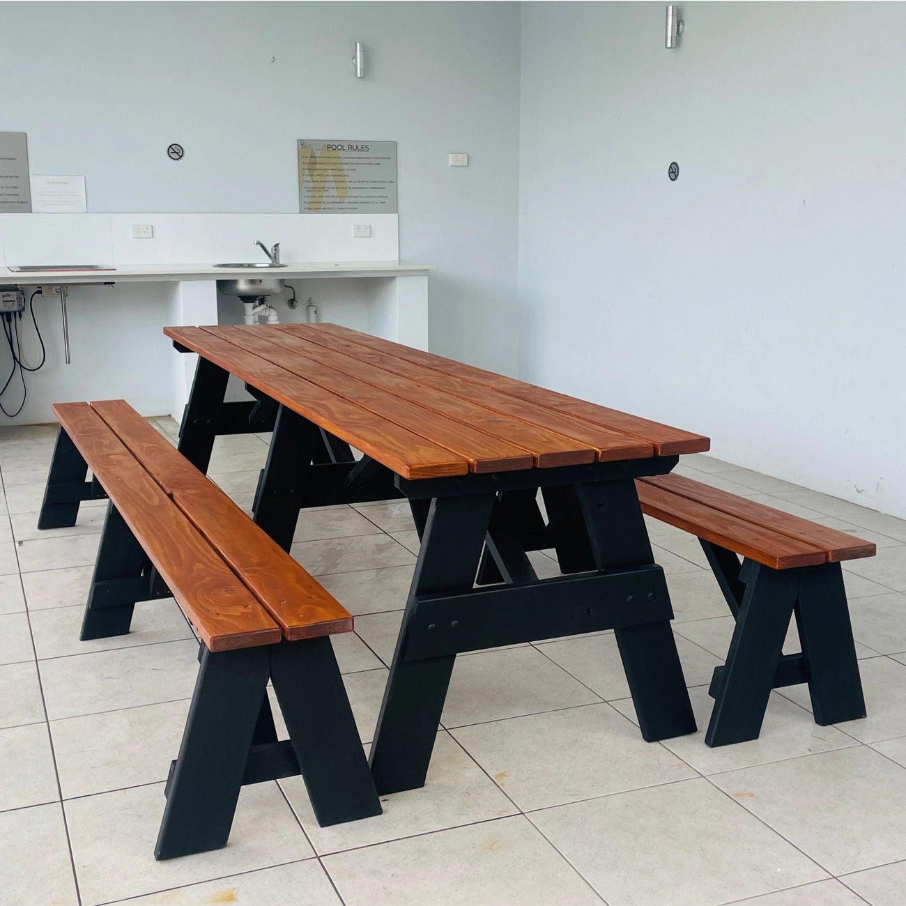 A wooden picnic table with two matching benches, featuring a warm brown wood top and black wooden A-frame legs, set in a tiled room with a white counter in the background.