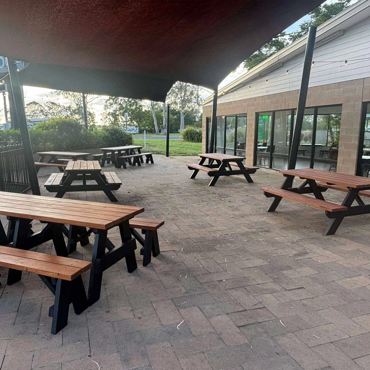 Outdoor patio seating area with multiple picnic tables and benches under a covered canopy, set on a paved patio in front of a modern building with large glass windows. Handmade by JJ Timber Design