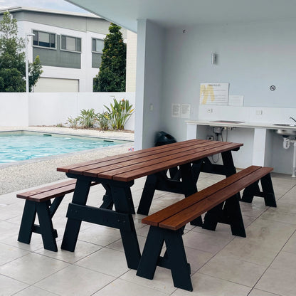 A wooden picnic table with two matching benches, featuring medium brown slatted tops and black wooden A-frame legs, set in an outdoor patio area next to a swimming pool.