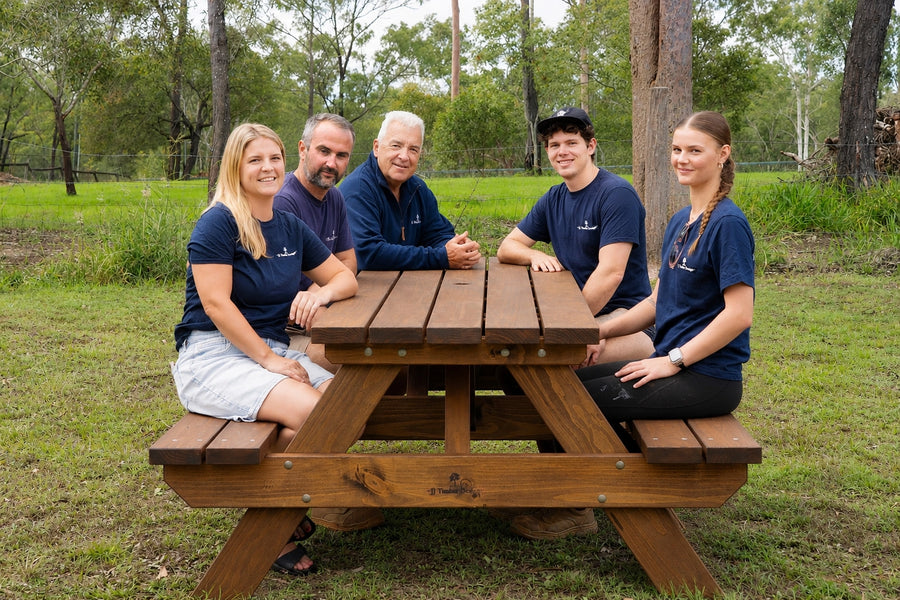 Five people wearing matching navy blue polo shirts sit together outdoors at a wooden picnic table on a grassy lawn with trees in the background.