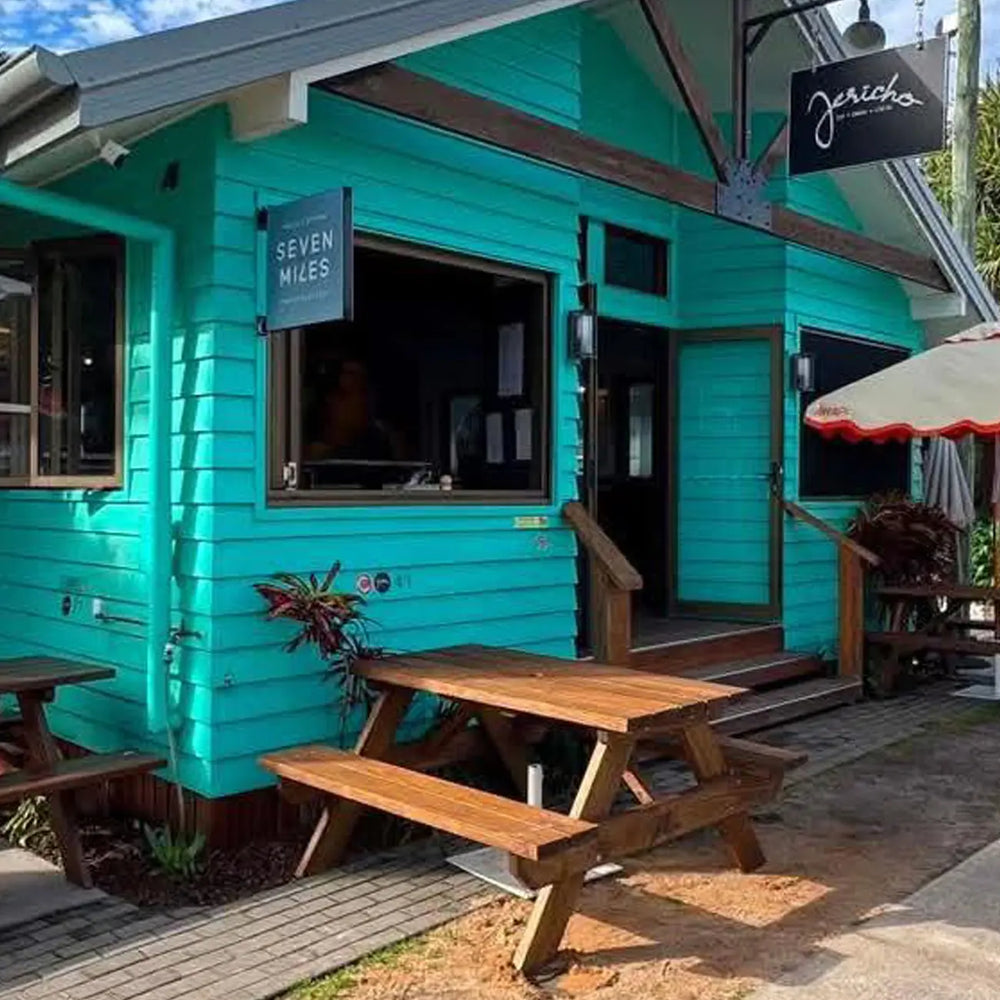 A bright turquoise wooden cafe building with outdoor picnic tables on a paved patio, a hanging black sign reading 'Seven Miles', and another hanging sign reading 'Jericho Cafe'.