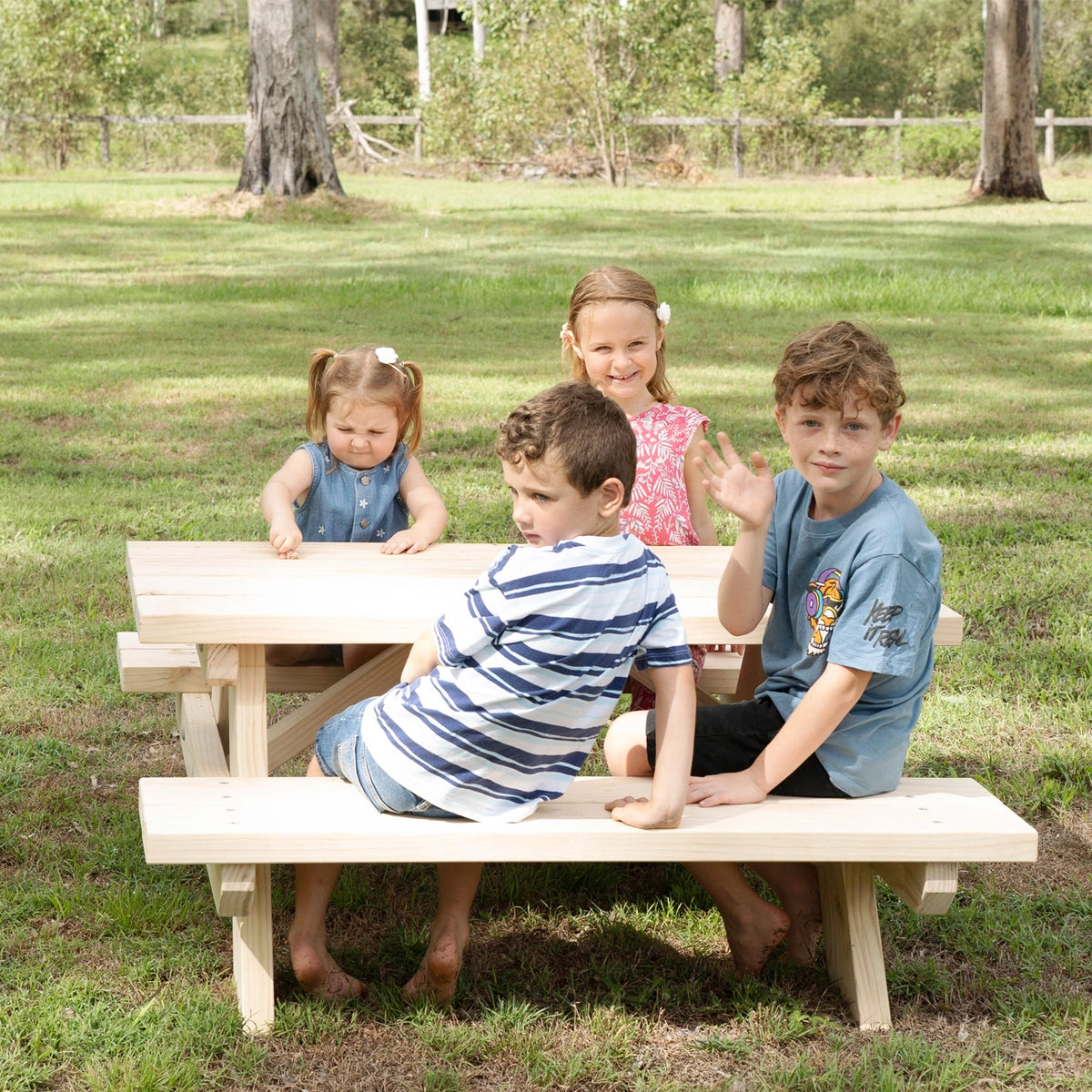 Four children sitting outdoors on a small wooden picnic table and bench in a grassy park setting.