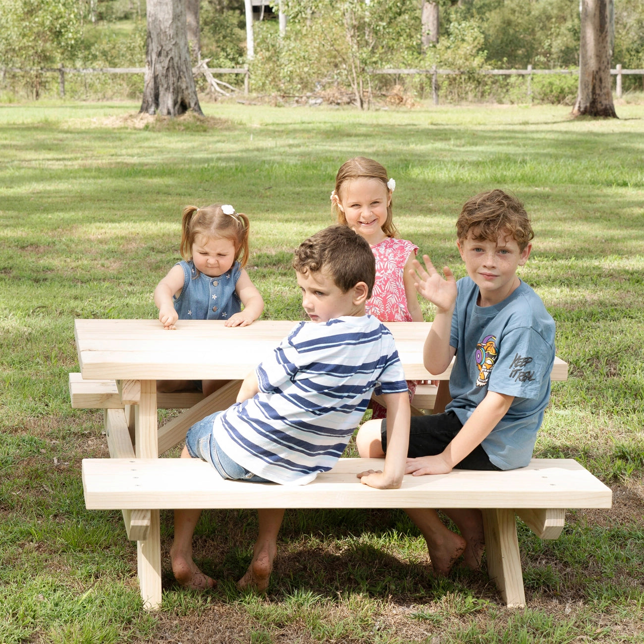 Four children sitting outdoors on a small wooden picnic table and bench in a grassy park setting.