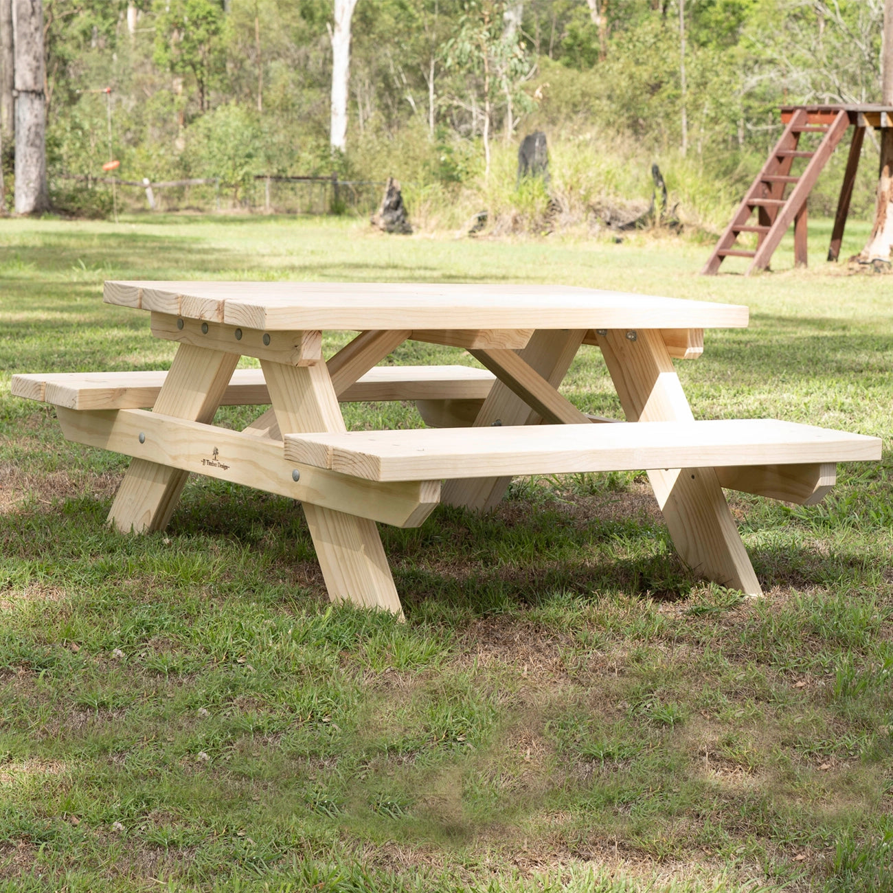A light natural wood picnic table with attached benches, shown outdoors on a grassy lawn with trees and a wooden play structure in the background. JJ Timber Design Hand Made
