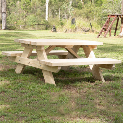 A light natural wood picnic table with attached benches, shown outdoors on a grassy lawn with trees and a wooden play structure in the background. JJ Timber Design Hand Made