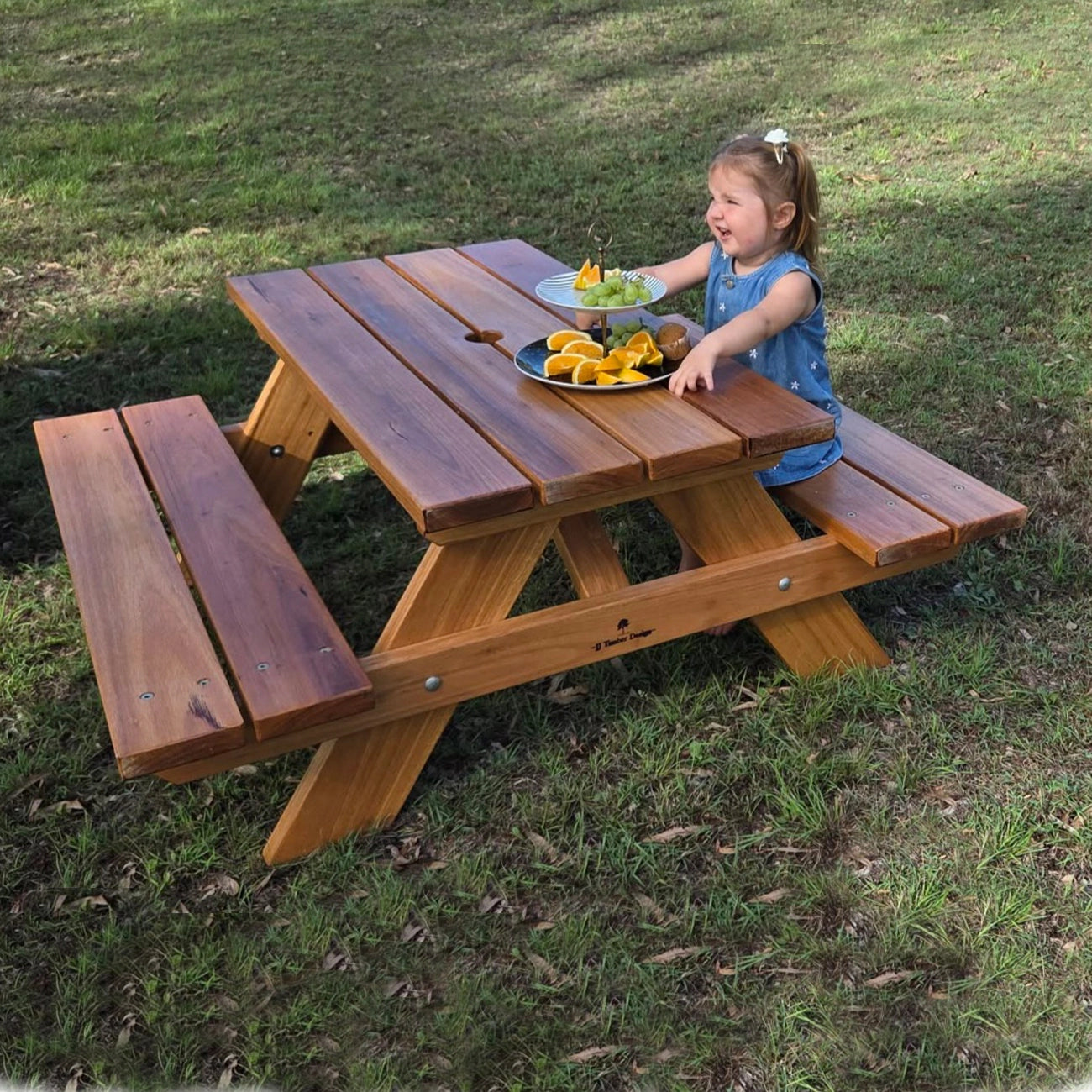 A young girl in a blue dress sits at a wooden picnic table outdoors on a grassy lawn, with plates of food on the table. Handmade by JJ Timber Design