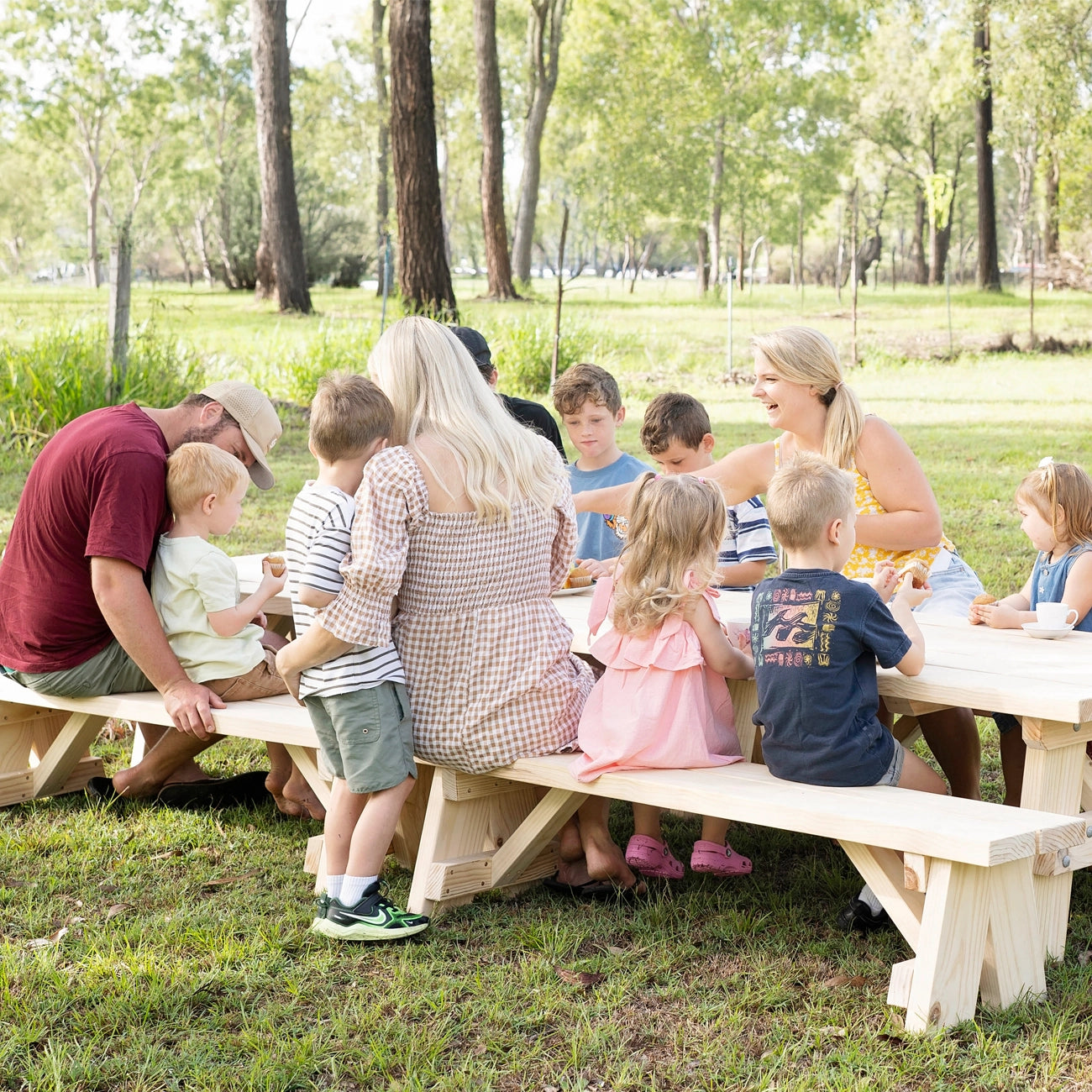 A group of adults and children gathered around a light natural wood picnic table outdoors in a grassy park with trees in the background.