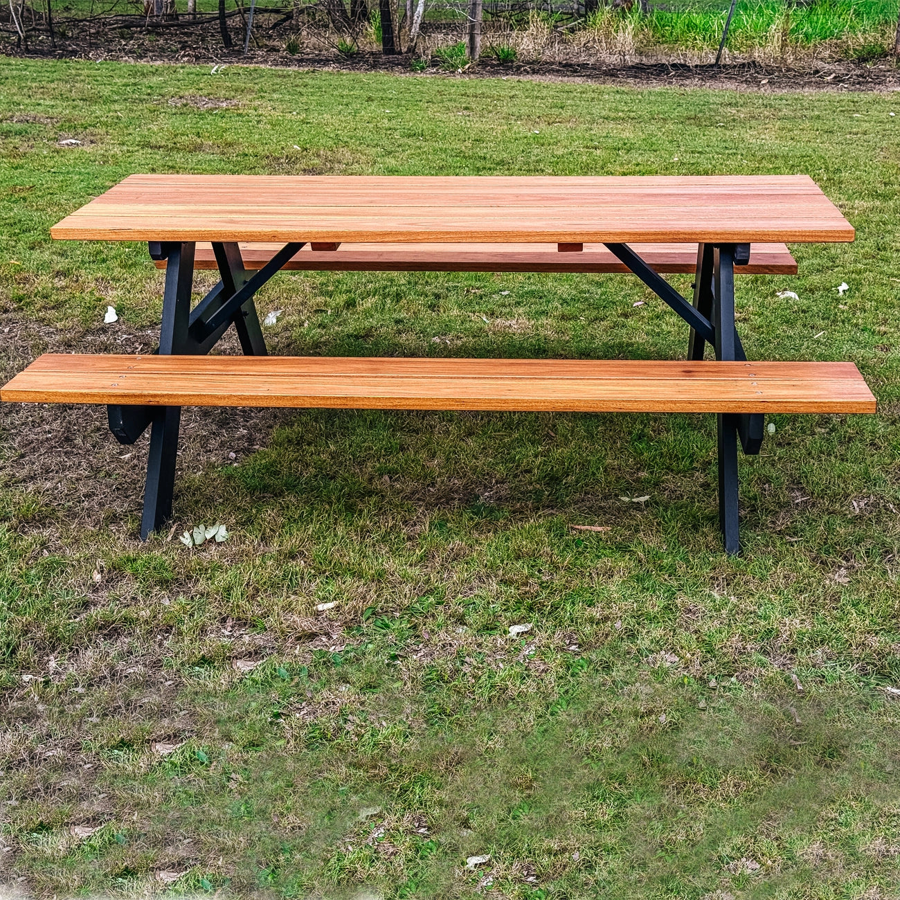 A wooden picnic table with attached benches, shown outdoors on a grassy lawn. The table has a warm medium-brown wood finish with black wooden support legs. Classic timber picnic table by JJ Timber Design installed on grass in Queensland