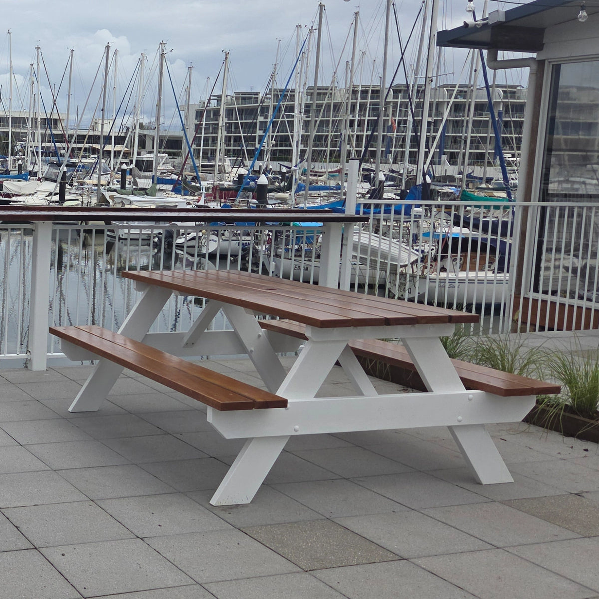 A white and brown wooden double picnic table on a tiled outdoor deck overlooking a marina with many boats and masts. Handmade by JJ Timber Design
