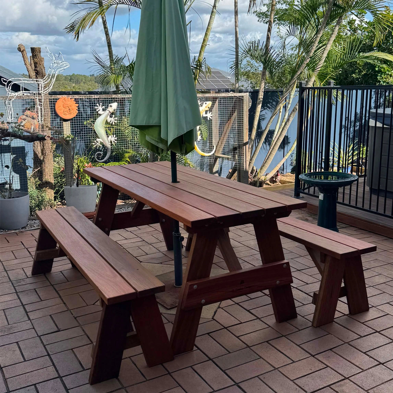 A dark brown wooden picnic table with two matching benches and a closed green patio umbrella, set on a brick patio with a lake and palm trees in the background. Handmade by JJ Timber Design