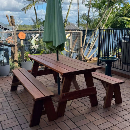 A dark brown wooden picnic table with two matching benches and a closed green patio umbrella, set on a brick patio with a lake and palm trees in the background. Handmade by JJ Timber Design