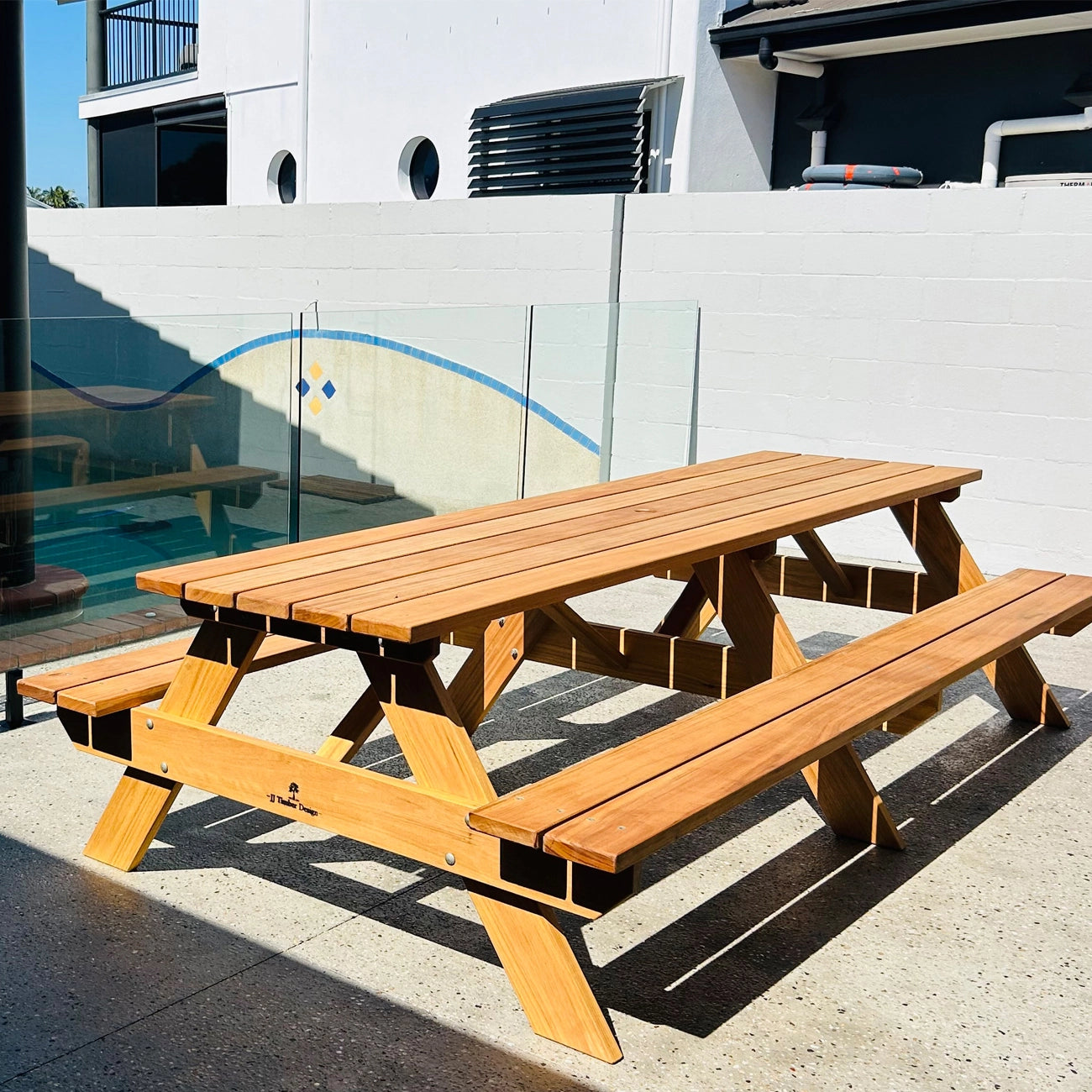 A wooden outdoor picnic table with attached benches, shown on a concrete patio in front of a white wall and glass railing. Hardwood Oiled Picnic Handmade by JJ Timber Design