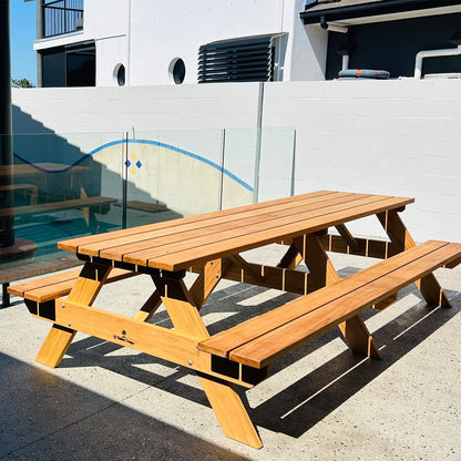 A wooden outdoor picnic table with attached benches, shown on a concrete patio in front of a white wall and glass railing. Hardwood Oiled Picnic Handmade by JJ Timber Design