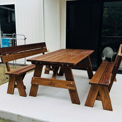 A dark brown wooden outdoor dining set consisting of a rectangular slatted table and two matching bench-style chairs, placed on a white patio in front of a building with white siding and a black door. Handmade by JJ Timber Design