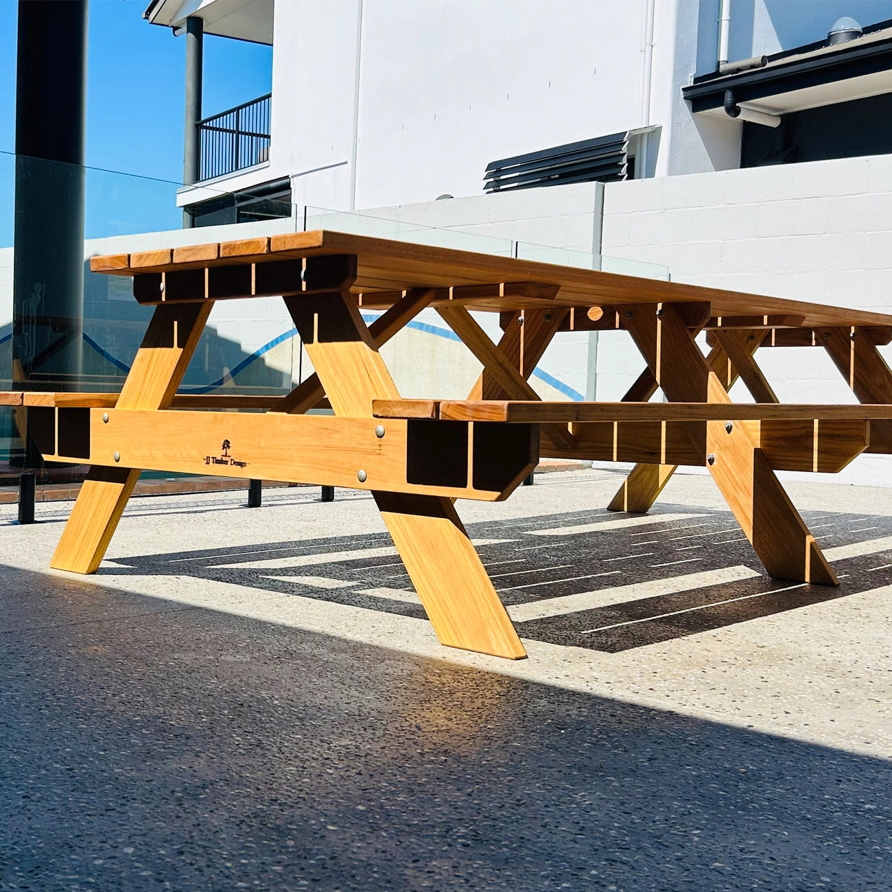 A light natural wood outdoor picnic table with integrated benches, photographed on a paved surface in front of a modern white building with glass windows. Handmade by JJ Timber Design