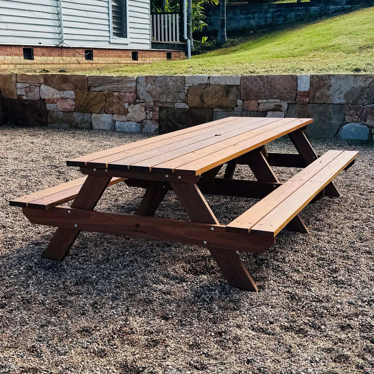A wooden picnic table with attached benches, shown outdoors on a gravel surface in front of a stone retaining wall and house siding. Handmade by JJ Timber Design