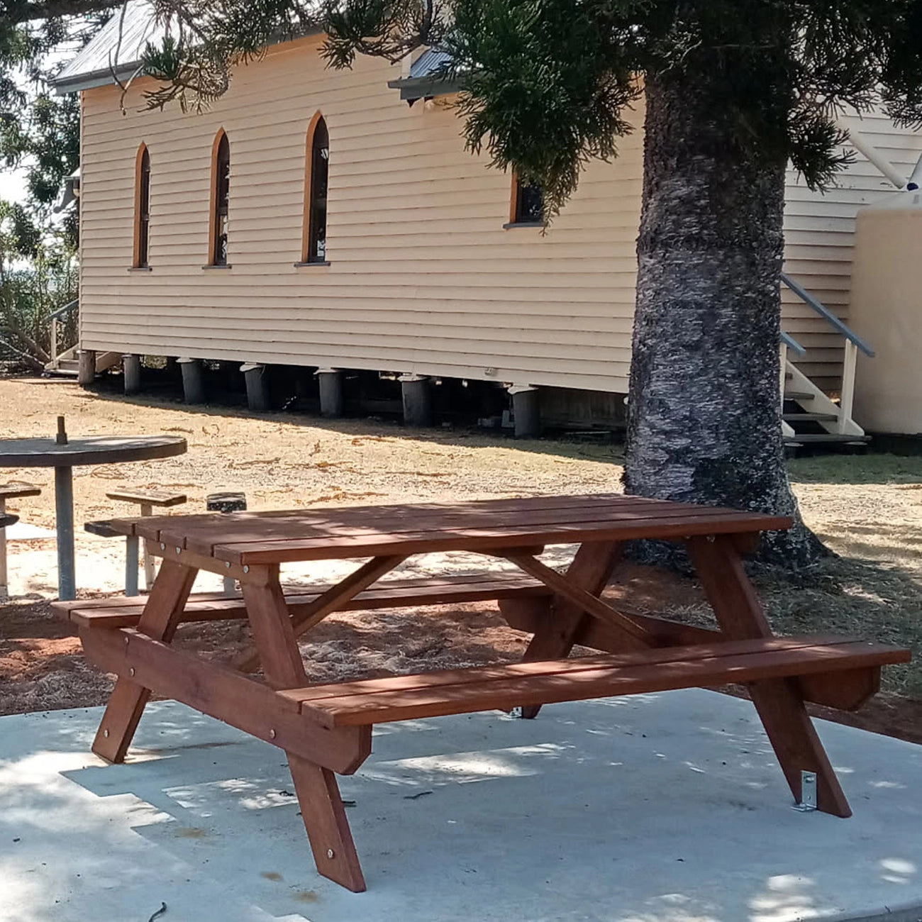 A reddish-brown wooden picnic table with attached benches is shown outdoors on a concrete patio, with a large tree trunk beside it and a beige building in the background. Handmade by JJ Timber Design
