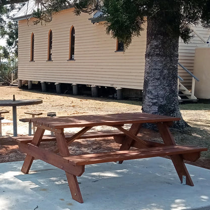 A reddish-brown wooden picnic table with attached benches is shown outdoors on a concrete patio, with a large tree trunk beside it and a beige building in the background. Handmade by JJ Timber Design