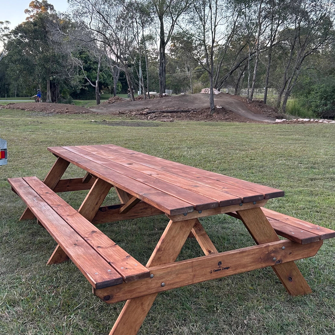 A rustic wooden picnic table with attached benches, shown outdoors on a grassy lawn with trees and a dirt track in the background. Handmade by JJ Timber Design