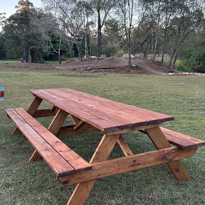 A rustic wooden picnic table with attached benches, shown outdoors on a grassy lawn with trees and a dirt track in the background. Handmade by JJ Timber Design