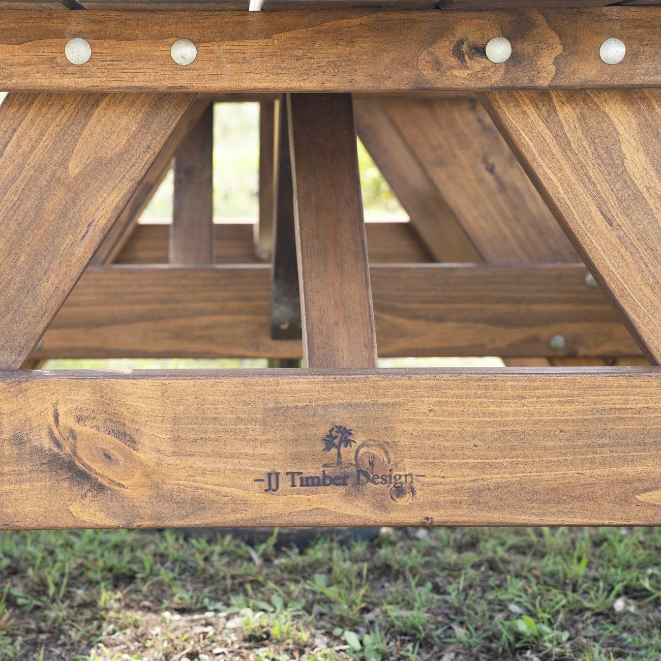 Close-up of a wooden picnic table frame outdoors, showing the engraved brand name 'JJ Timber Design' on the front rail above grass. Handmade by JJ Timber Design