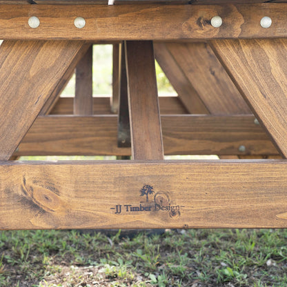 Close-up of a wooden picnic table frame outdoors, showing the engraved brand name 'JJ Timber Design' on the front rail above grass. Handmade by JJ Timber Design