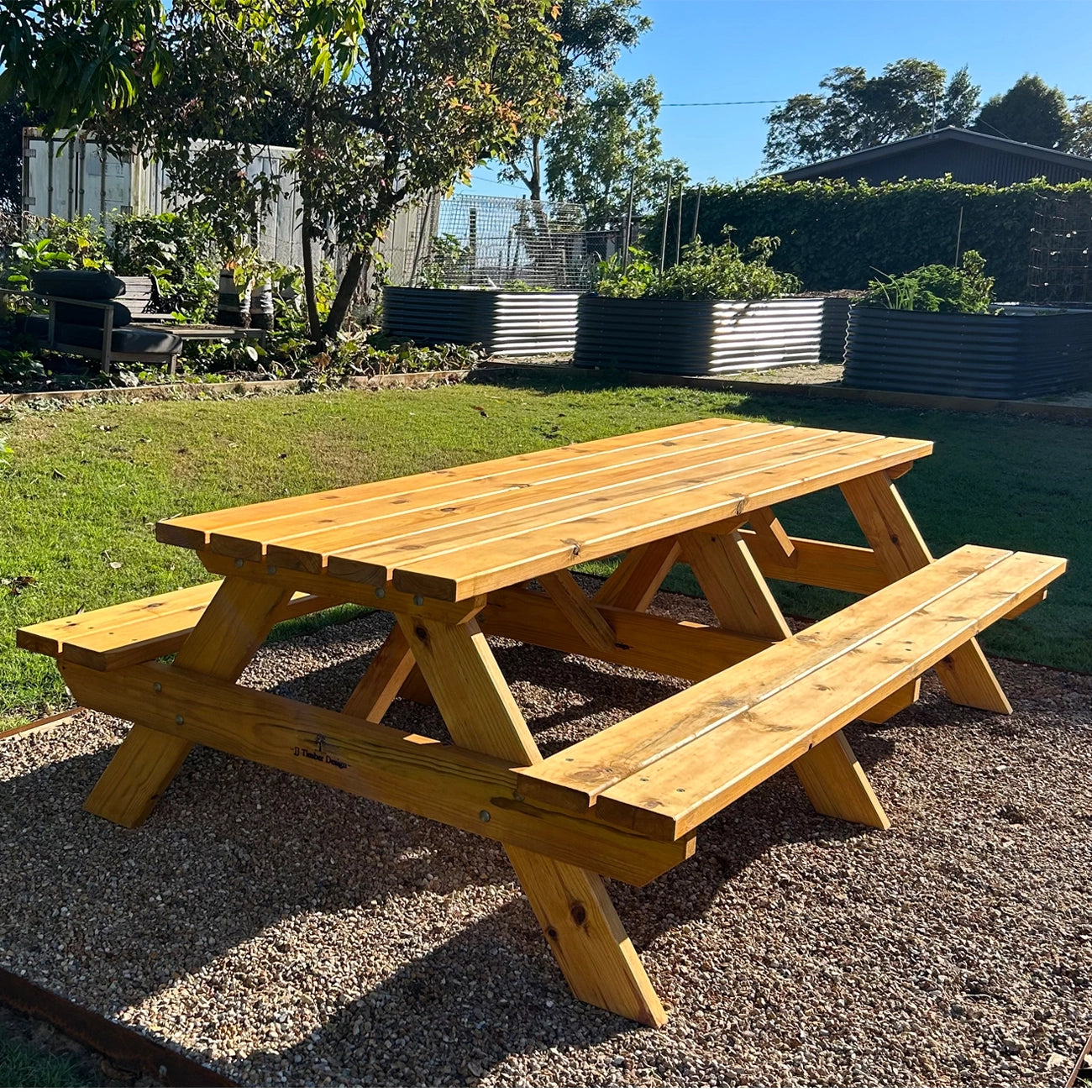 A wooden outdoor picnic table with attached benches, shown in a backyard setting on a gravel surface with grass, trees, and planter boxes in the background. Handmade by JJ Timber Design