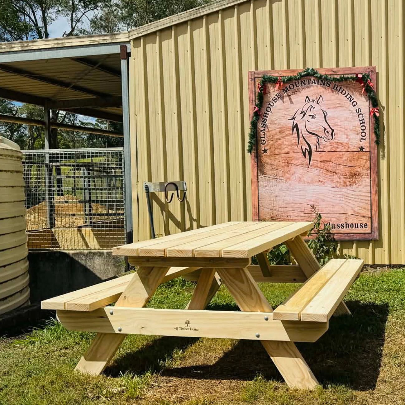 A light natural wood picnic table with attached benches is set outdoors in front of a corrugated metal building. A large wooden sign on the wall reads Classhouse Mountain Riding School, and the table has a small Classhouse logo printed on the front apron. Handmade by JJ Timber Design