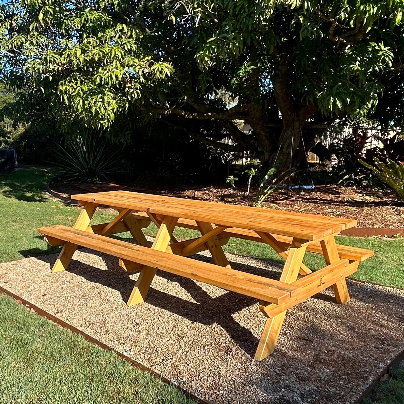 A wooden picnic table with attached benches, shown outdoors on a gravel pad with grass, trees, and garden plants in the background. Handmade by JJ Timber Design