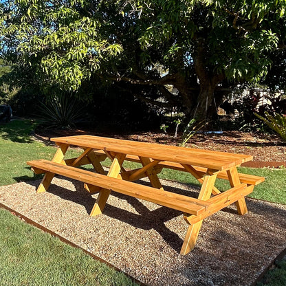 A wooden picnic table with attached benches, shown outdoors on a gravel pad with grass, trees, and garden plants in the background. Handmade by JJ Timber Design
