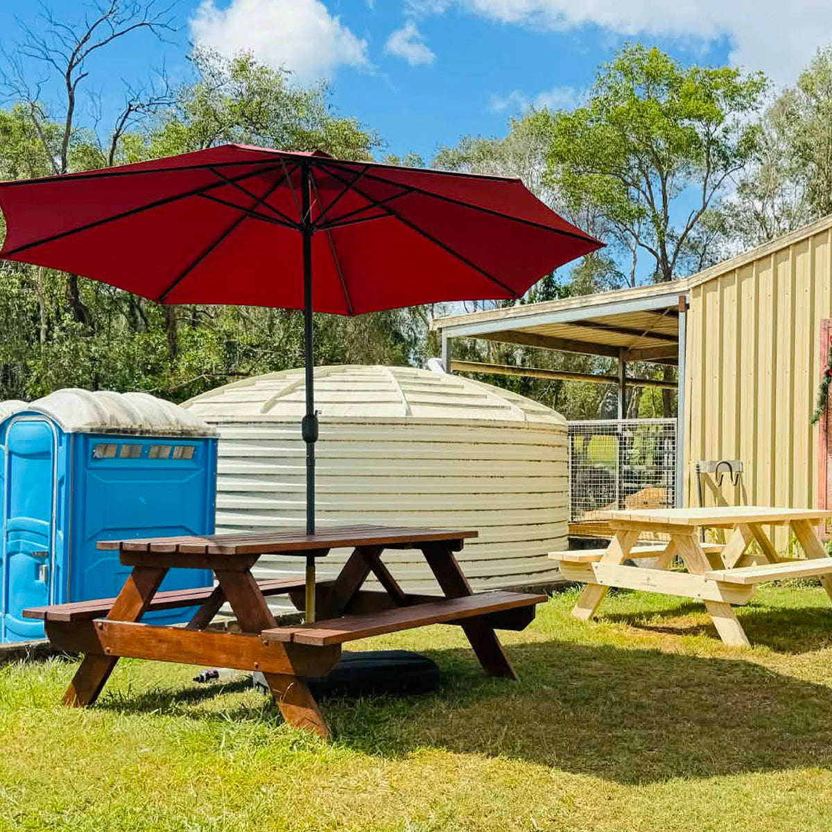 Outdoor backyard scene with a large red patio umbrella over a wooden picnic table, a white corrugated storage container, a blue portable toilet, another picnic table, and a beige shed under a blue sky with trees. Handmade by JJ Timber Design