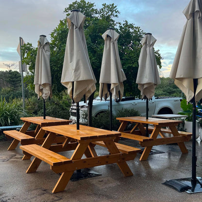 Outdoor wooden picnic tables with attached benches arranged on a patio, with closed beige patio umbrellas and a white pickup truck in the background. Handmade by JJ Timber Design