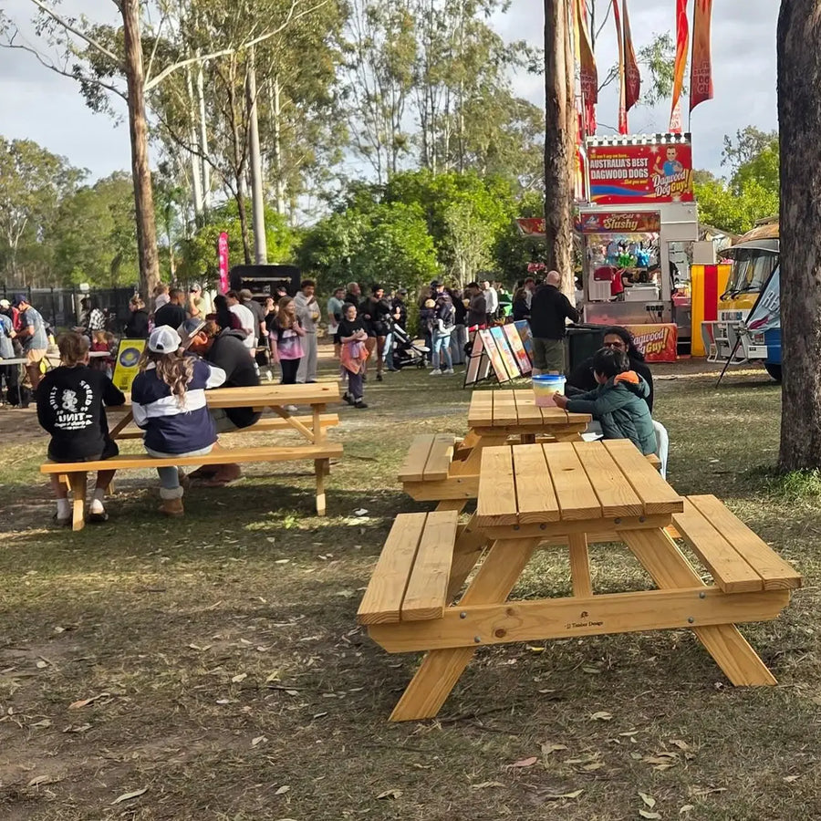 Outdoor park scene with several light natural wood picnic tables in the foreground, people sitting and walking near food trucks and trees in the background.