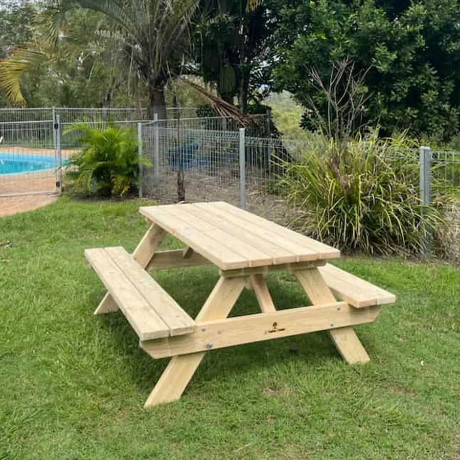 A light natural wood picnic table with attached benches is set on a green grass lawn in an outdoor backyard setting with a pool, fence, and trees in the background.
