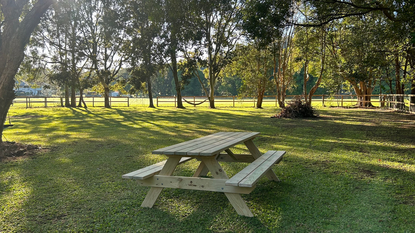 A light-colored wooden picnic table with attached benches sits on a grassy lawn in a park with trees and a fence in the background.