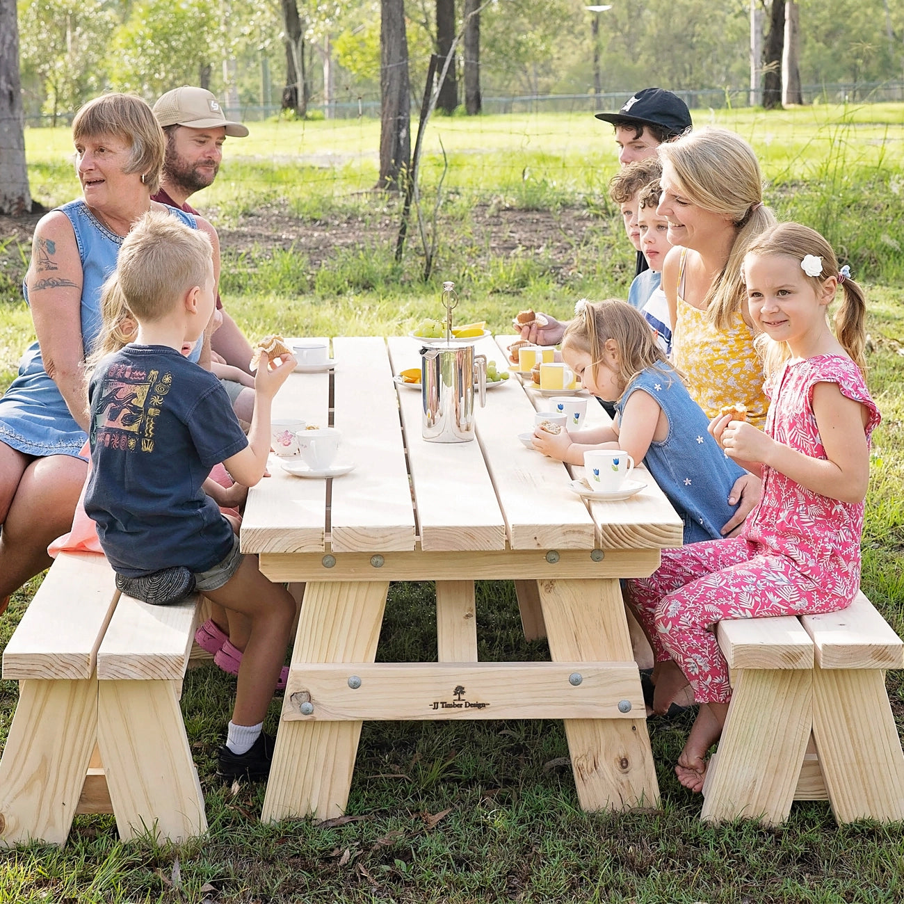 A family of six gathered around a light natural wood picnic table with matching benches in a grassy park setting. The table features a slatted top and thick block legs, with a small 'The Picnic Place' logo printed on the front center support.
