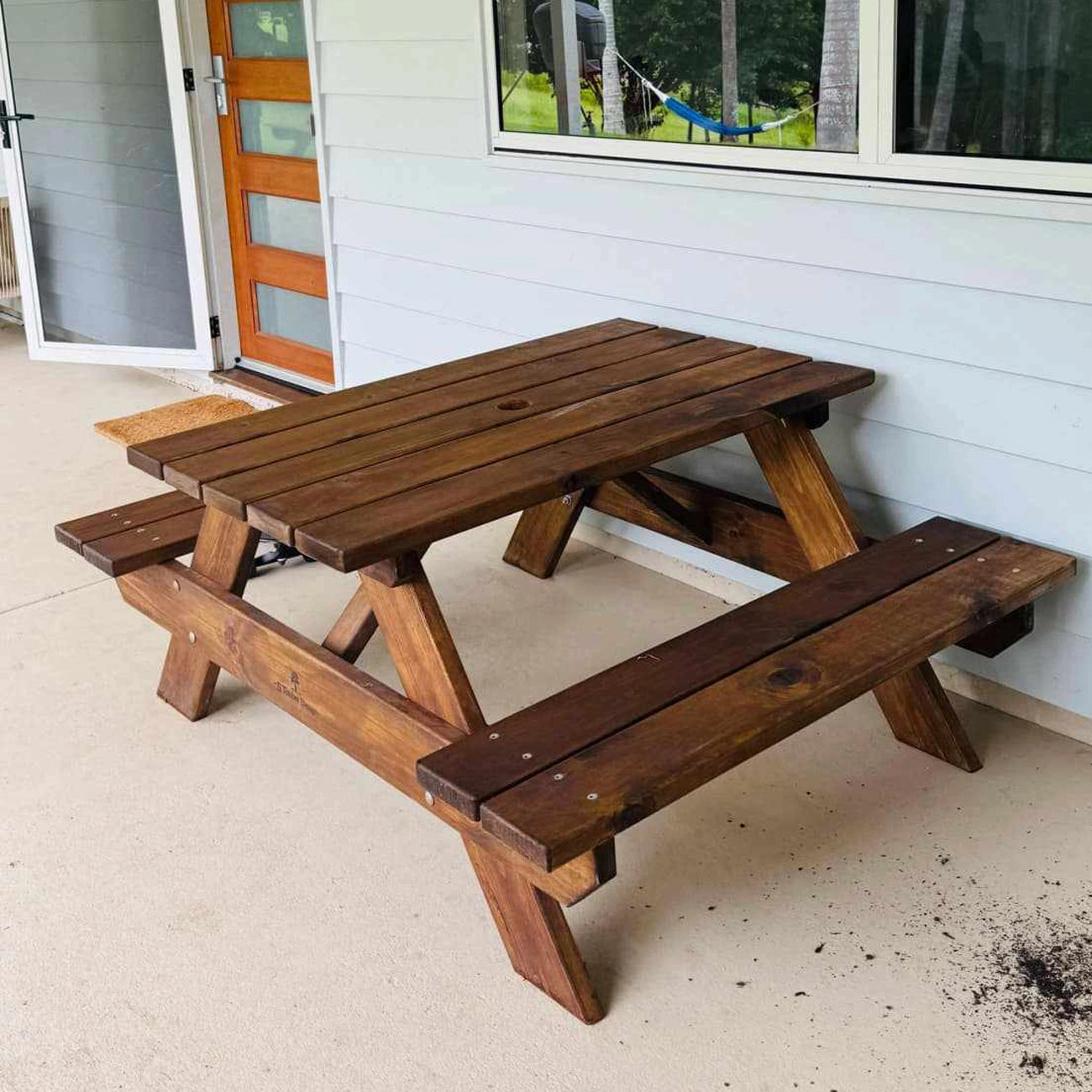 A dark brown wooden picnic table with attached benches, shown outdoors on a concrete patio against a white-sided house. JJ Timber Design Picnic Table