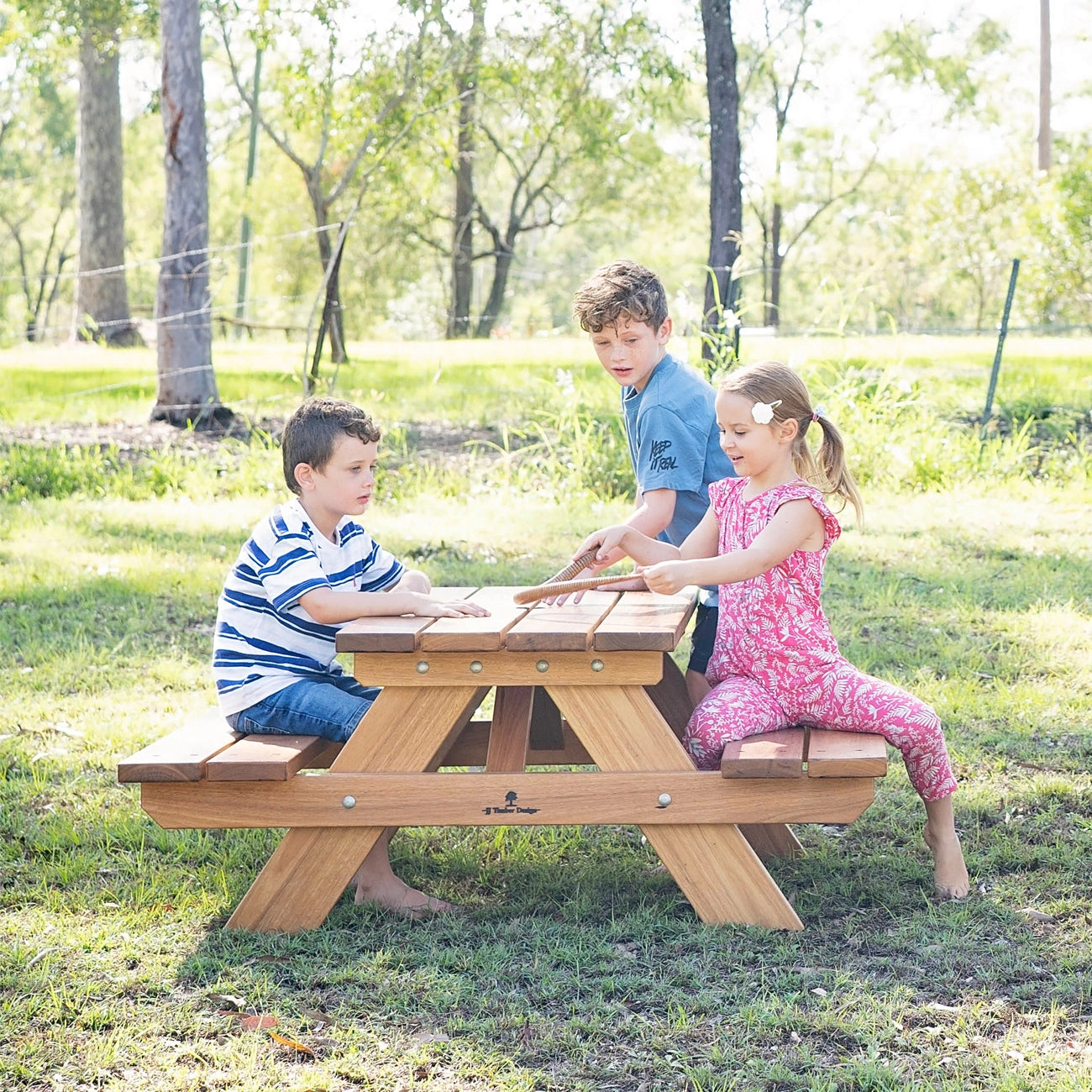 A wooden outdoor picnic table with three children sitting and playing on a grassy lawn in a park setting.