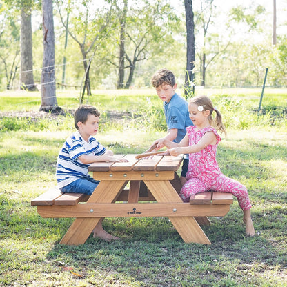 A wooden outdoor picnic table with three children sitting and playing on a grassy lawn in a park setting.