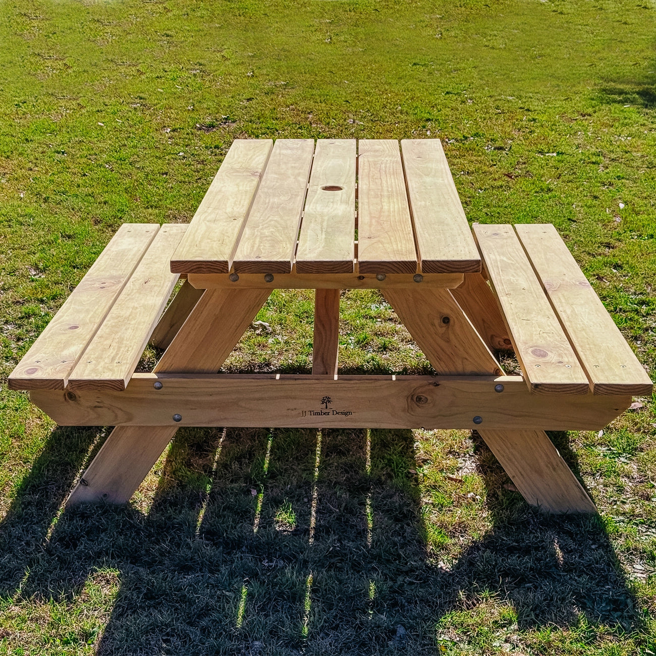 A light natural wood picnic table with attached benches, photographed outdoors on a grassy lawn in bright sunlight. Handmade by JJ Timber Design