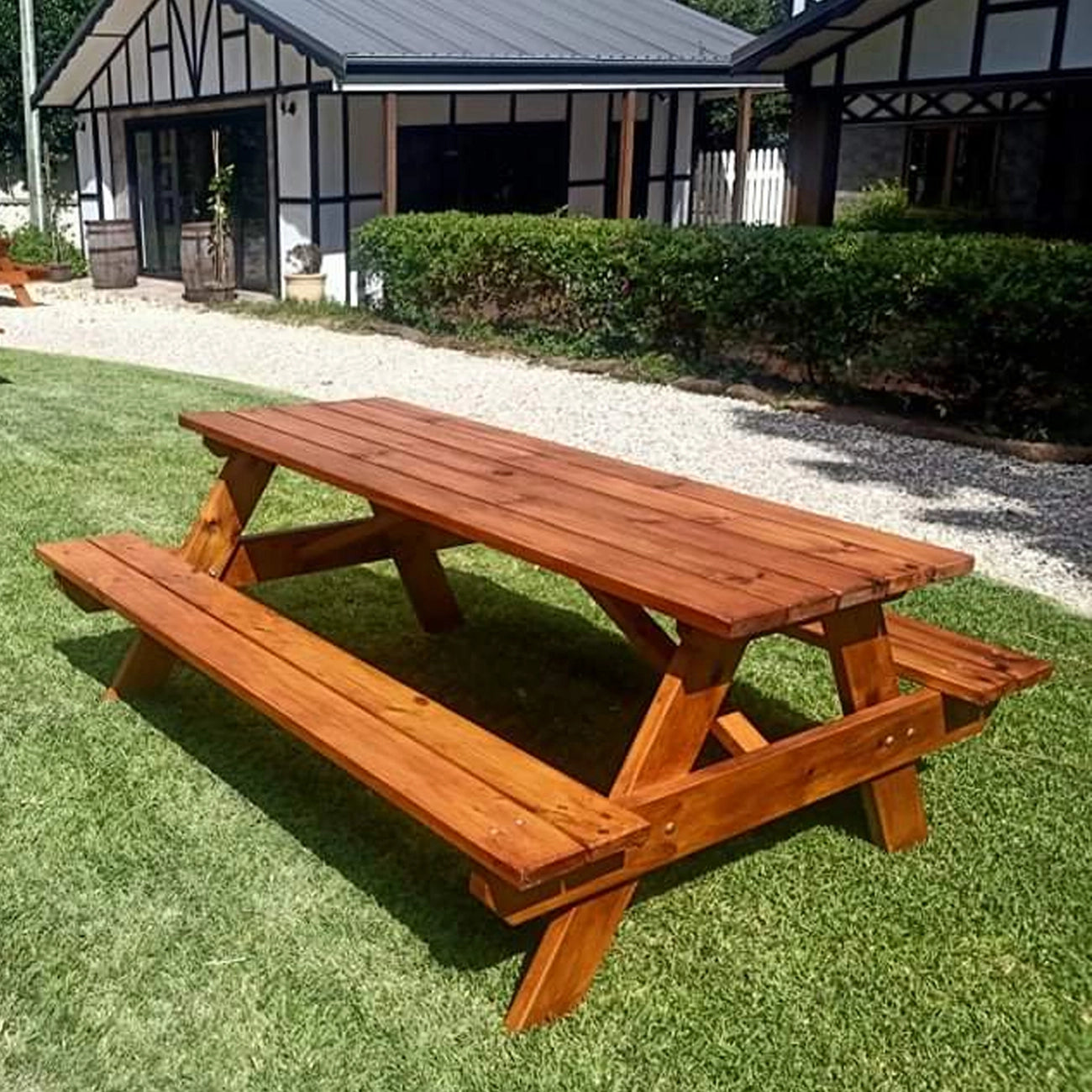 A wooden picnic table with attached benches, shown outdoors on a green lawn in front of a house with a gravel path and hedges. Handmade by JJ Timber Design