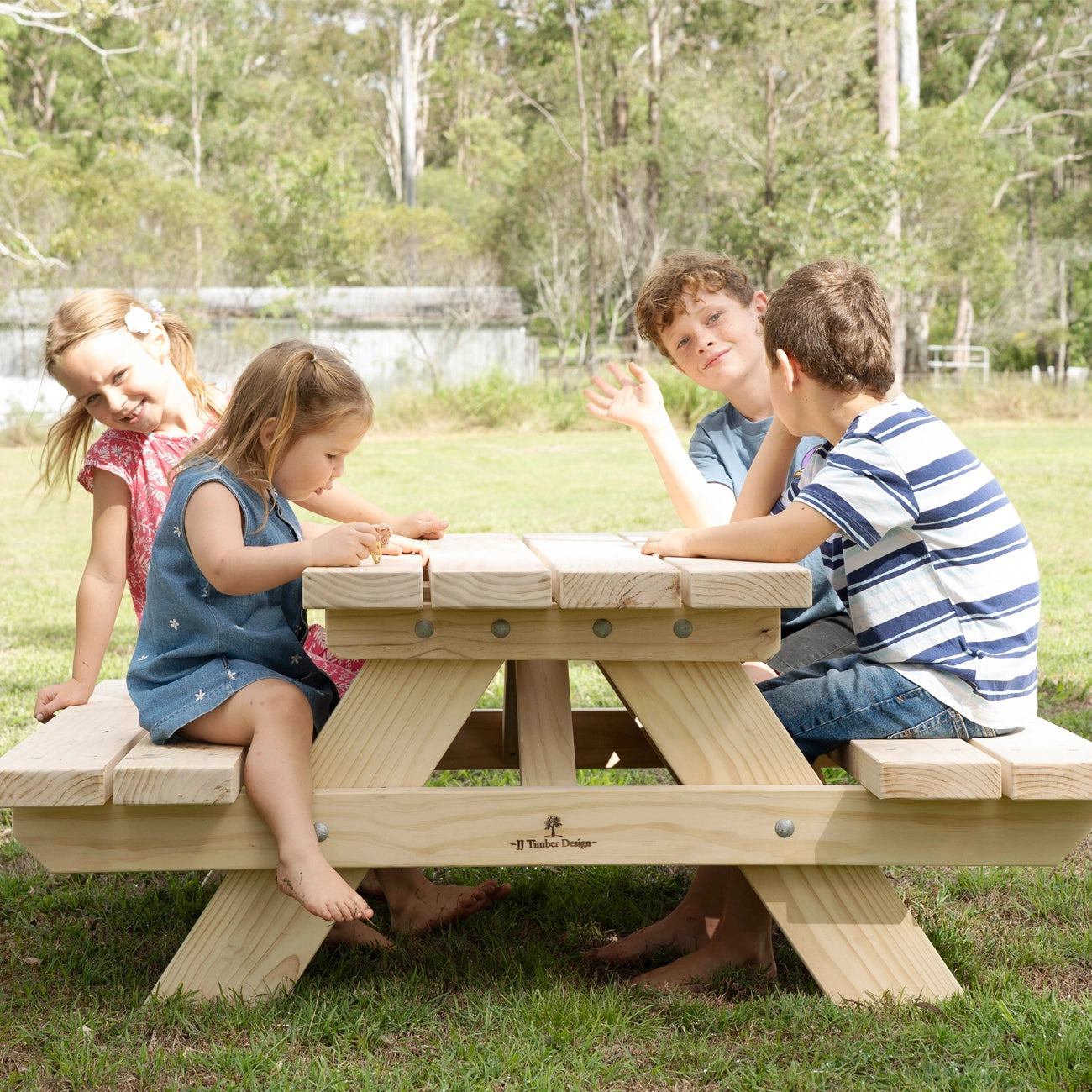 A children's wooden picnic table with built-in benches is shown outdoors in a grassy park setting, with four children sitting and playing around it.