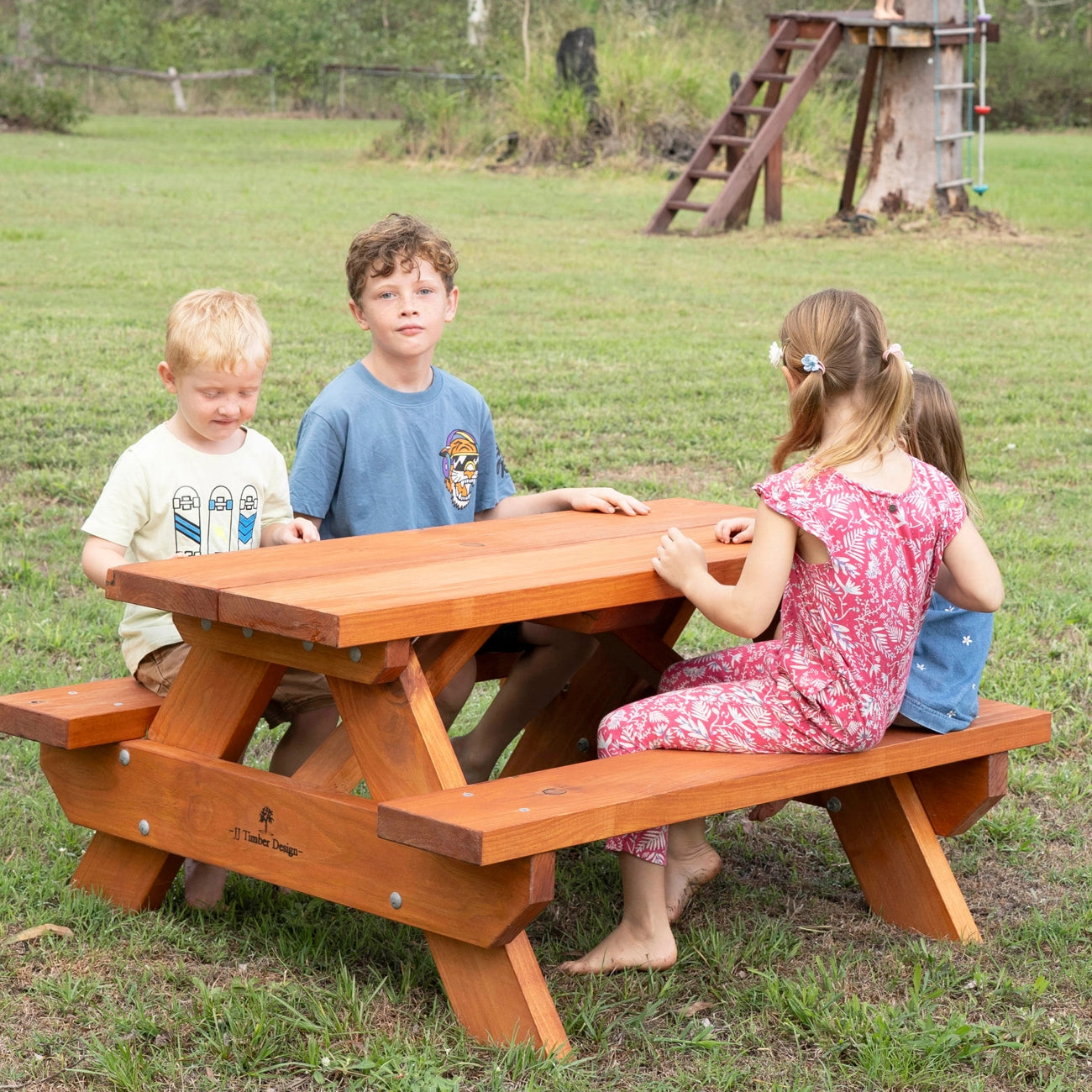 A children's wooden picnic table with attached benches is shown outdoors on grass, with three children sitting and standing around it. A treehouse play structure is visible in the background.
