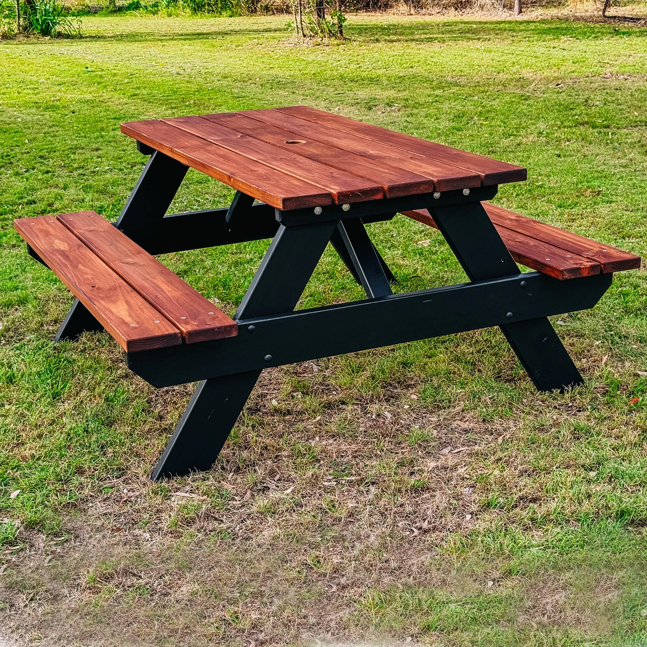 A wooden picnic table with attached benches, featuring a reddish-brown stained wood top and black wooden frame, set on a grassy lawn. Handmade by JJ Timber Design